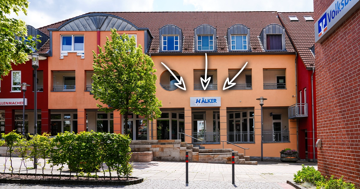 Ein mehrstöckiges Gebäude in warmen Erdtönen mit "HÄLKER"-Schild, umgeben von Grünpflanzen.
