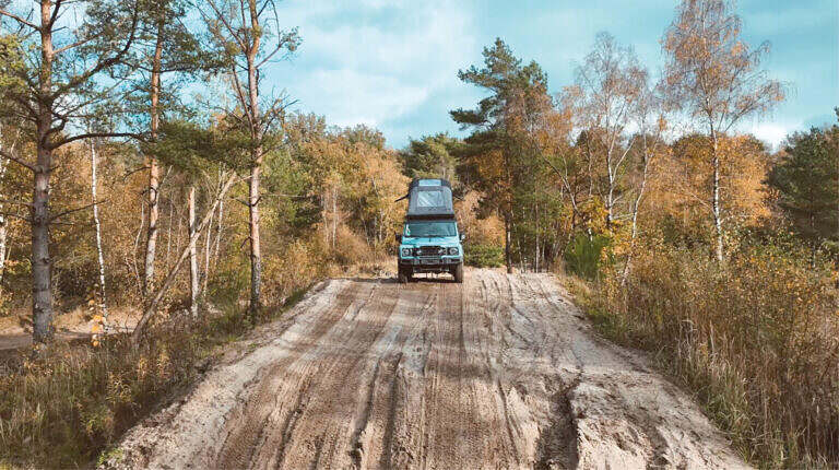 Ein hellblauer Geländewagen mit Dachzelt steht auf einem sandigen Waldweg im herbstlichen Laubwald.