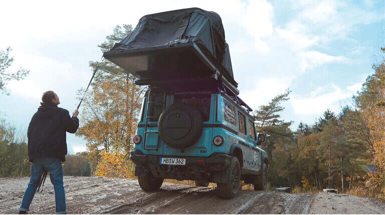 Ein türkisfarbener Geländewagen mit Dachzelt in herbstlicher Natur, Person richtet das Zelt auf.