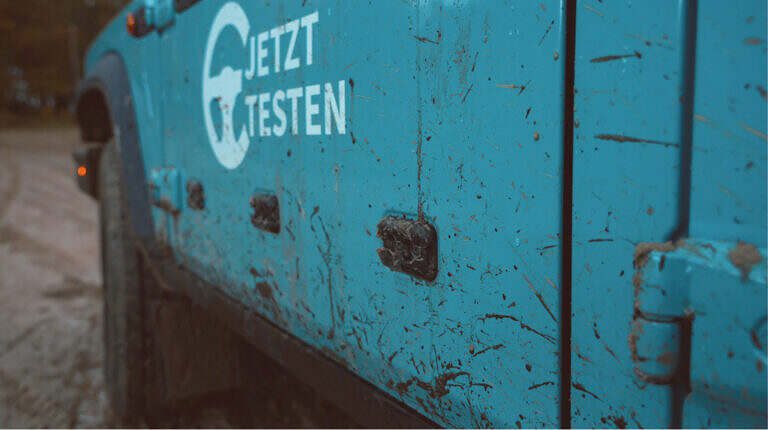 Seitliche Ansicht eines blauen, schlammigen Geländewagens mit dem Schriftzug "JETZT TESTEN" auf der Tür.