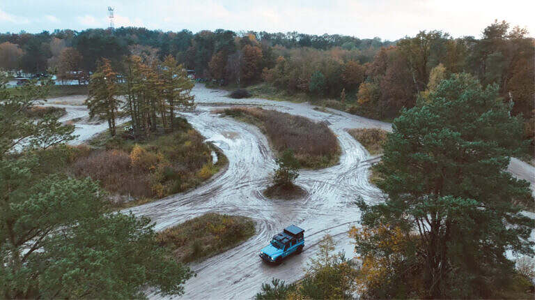Blaues Offroad-Fahrzeug auf kurvigem, sandigem Waldweg, umgeben von herbstlicher Vegetation.