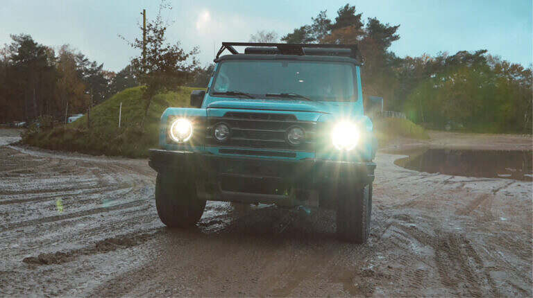 Ein blaues Geländefahrzeug fährt mit leuchtenden Scheinwerfern auf einem schlammigen Weg durch die Natur.