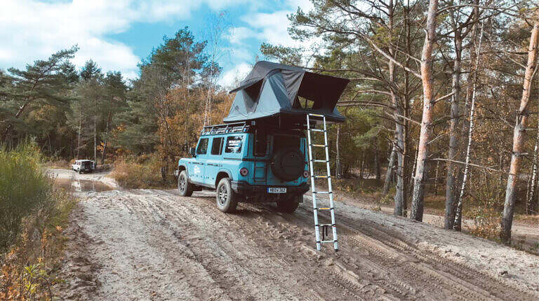 Blaues Offroad-Fahrzeug mit Dachzelt auf sandigem Weg im Wald, herbstliche Atmosphäre.