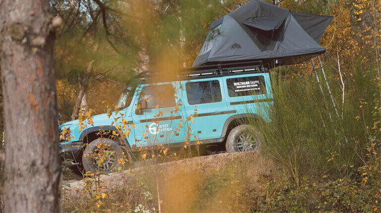 Türkisfarbenes Geländefahrzeug mit Dachzelt, geparkt in einem herbstlichen Wald. Abenteuerliche Stimmung.