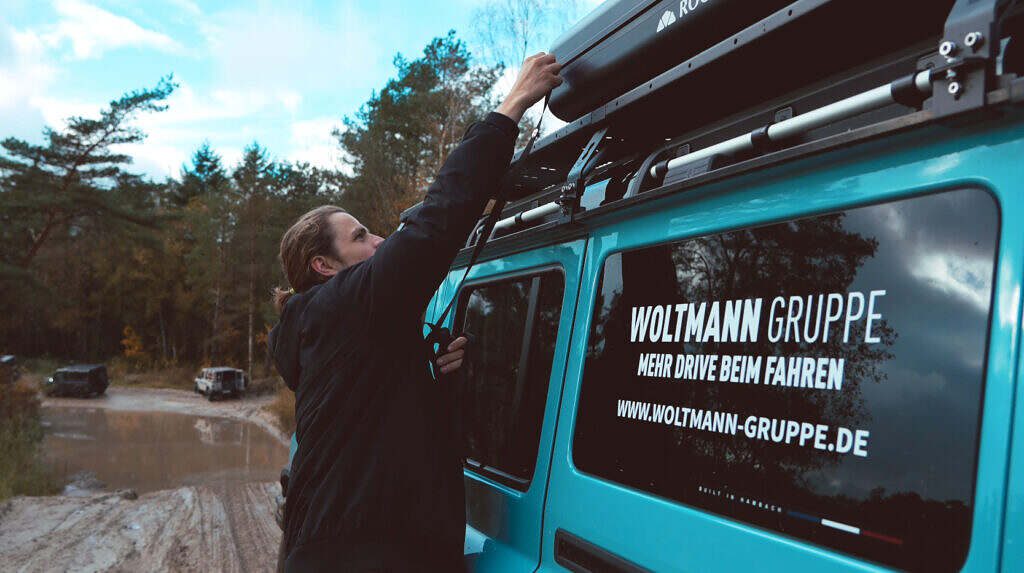 Blauer Geländewagen im Matsch, Mann befestigt Gepäck auf dem Dach, umgeben von Bäumen und Pfützen.