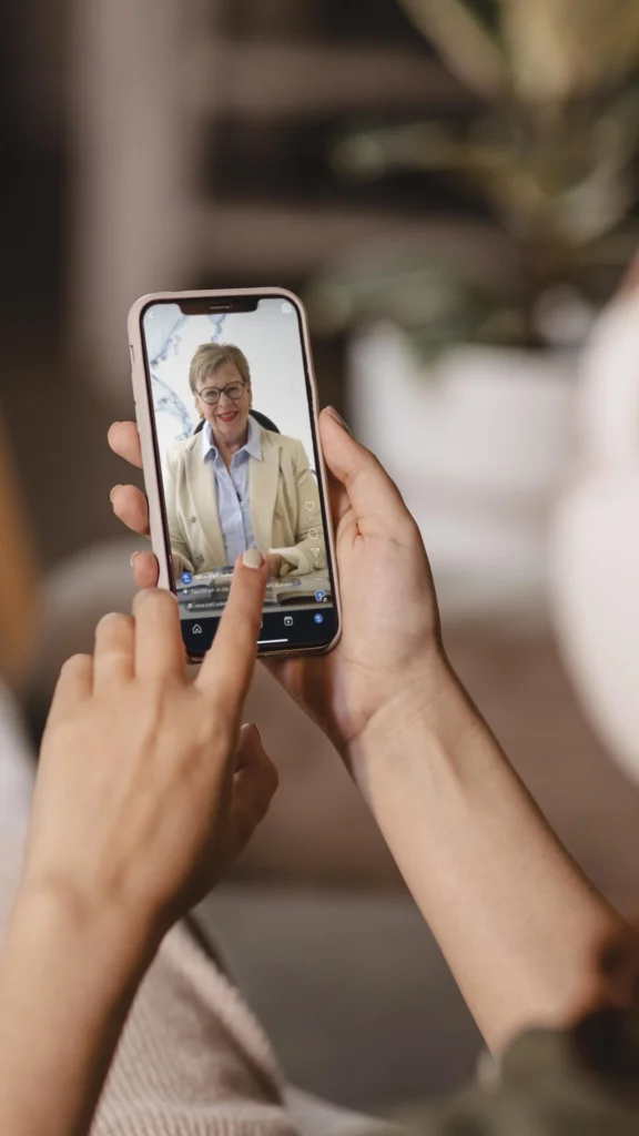 Hände halten ein Smartphone mit einem Videoanruf, der eine lächelnde Person im hellen Sakko zeigt.