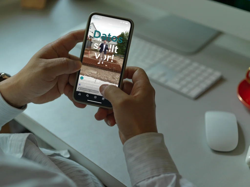Person hält Smartphone mit Social-Media-App, Schreibtisch mit Tastatur und Monitor im Hintergrund.