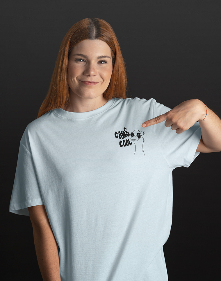 Eine lächelnde Person mit rötlichem Haar zeigt auf die Grafik "CANS COOL" auf ihrem hellblauen T-Shirt.