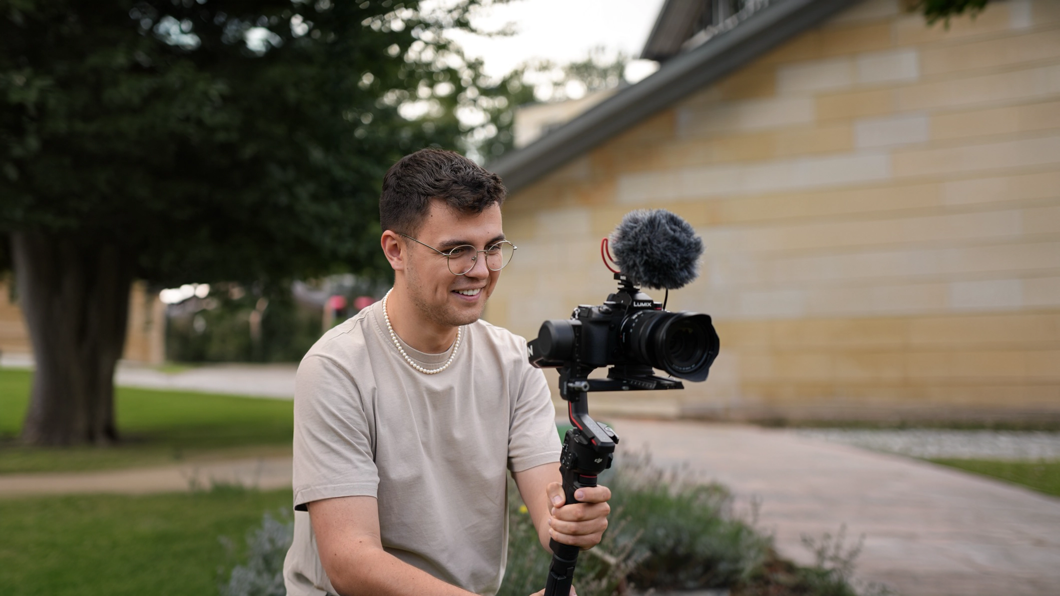 Person mit Brille hält Kamera auf Gimbal, filmt draußen vor grüner Kulisse, wirkt konzentriert und entspannt.