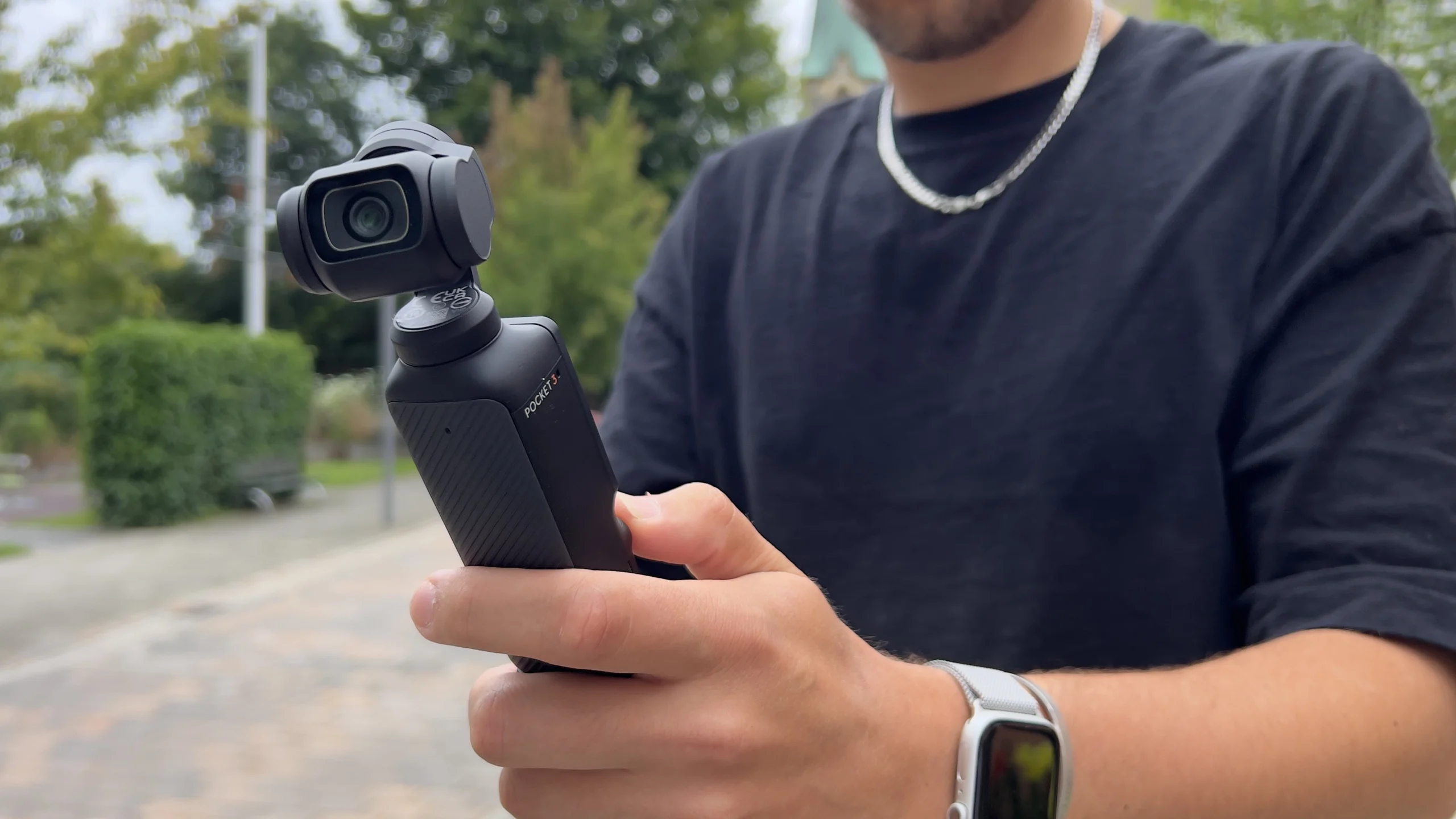 Person hält kompakte Kamera mit Gimbal, bereit für Aufnahme. Hintergrund unscharf, grün-graue Töne.