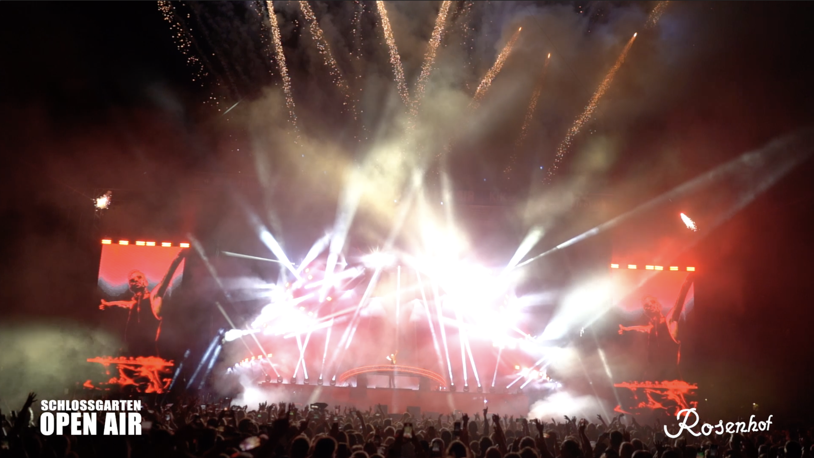 Ein DJ animiert eine jubelnde Menge bei einem Open-Air-Konzert mit beeindruckender Lichtshow und Feuerwerk.