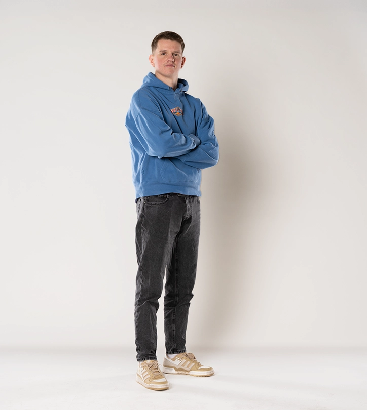 Eine Person in blauem Hoodie und dunklen Jeans steht entspannt mit verschränkten Armen vor weißem Hintergrund.