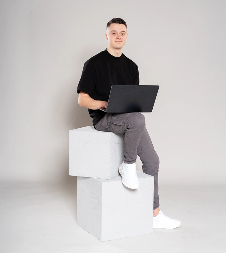 Person sitzt auf weißen Würfeln, arbeitet am Laptop; trägt schwarzes T-Shirt, graue Hose, weiße Schuhe.