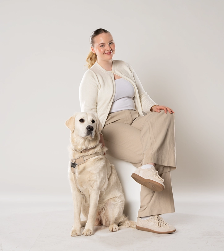 Eine lächelnde Frau sitzt auf einem Hocker, streichelt ihren großen, cremefarbenen Hund.