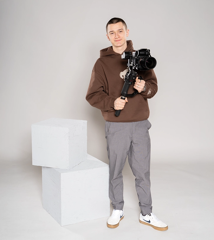 Person mit Kamera auf Stabilisator, lächelt freundlich, steht neben zwei großen weißen Würfeln.