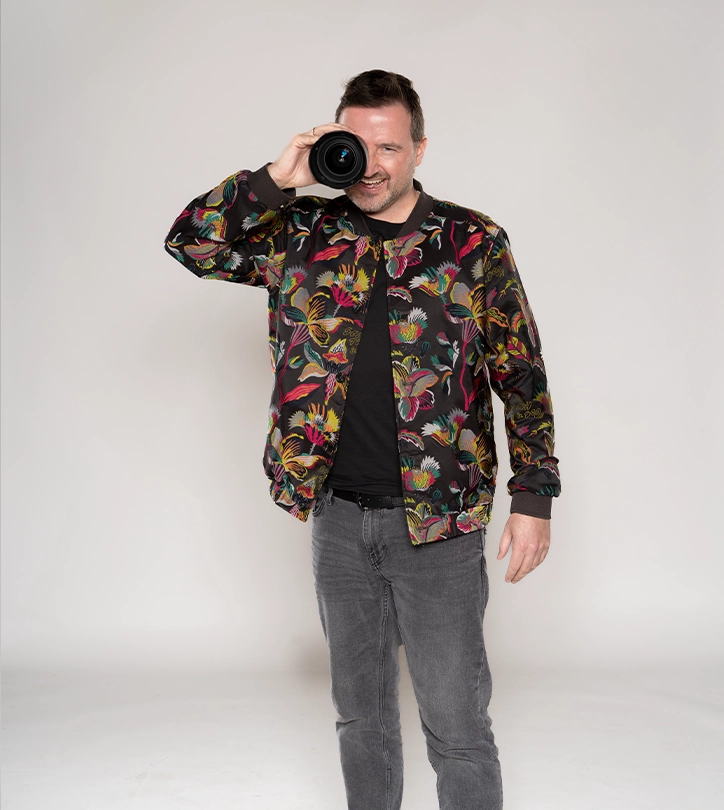 Eine lächelnde Person hält ein Kameraobjektiv vors Auge, trägt eine bunte Jacke vor hellem Hintergrund.