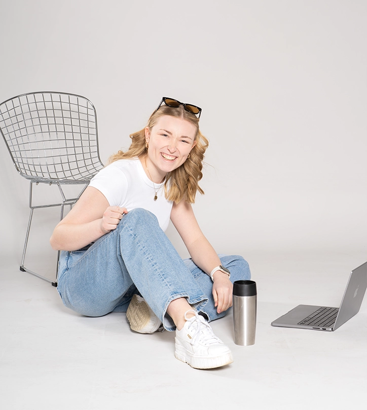 Junge Frau sitzt lächelnd auf dem Boden mit Laptop und Thermobecher, in entspannter, moderner Umgebung.