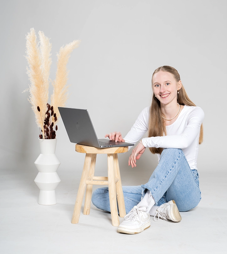 Junge Frau sitzt lächelnd am Boden, arbeitet am Laptop, umgeben von minimalistischer Einrichtung.