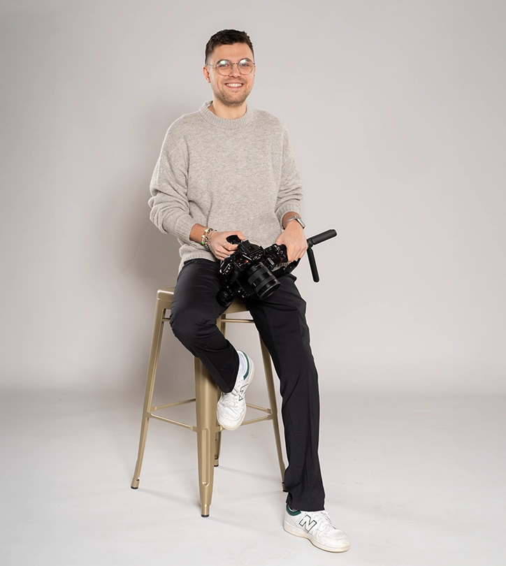 Person auf Hocker mit Kamera, lächelnd, in grauem Pullover und schwarzen Hosen vor grauem Hintergrund.