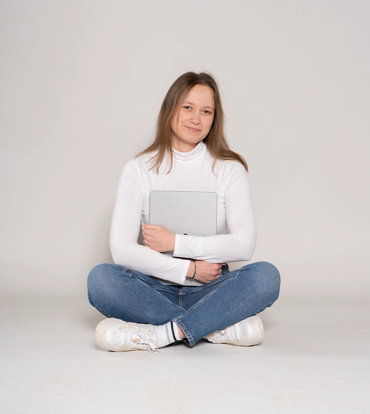 Person im Schneidersitz hält Laptop, trägt weißes Oberteil, blaue Jeans, weiße Turnschuhe, lächelt.