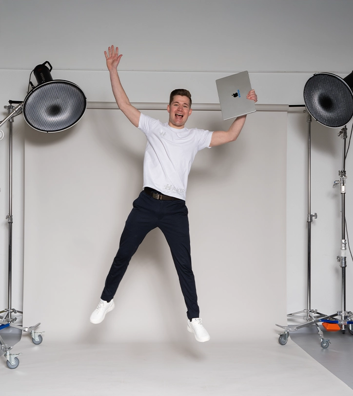 Person springt freudig im Fotostudio, hält Laptop in der Hand, umgeben von Studiolampen.
