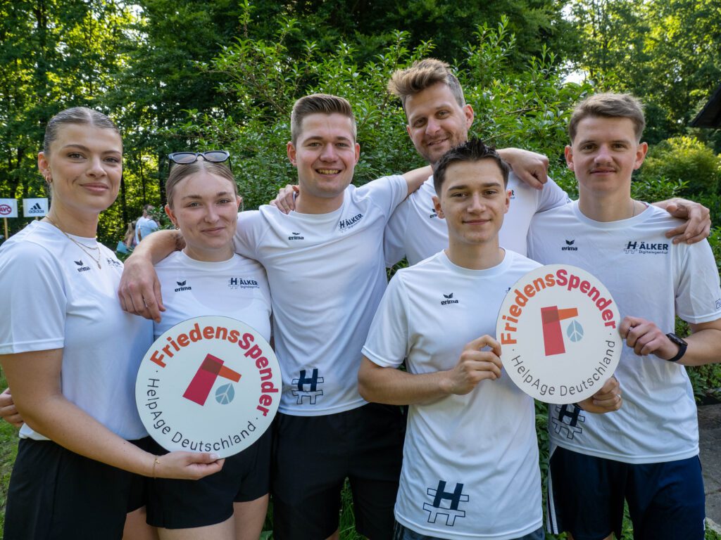 Sechs Personen in weißen Sportshirts halten Schilder von "FriedensSpender - HelpAge Deutschland" im Park.
