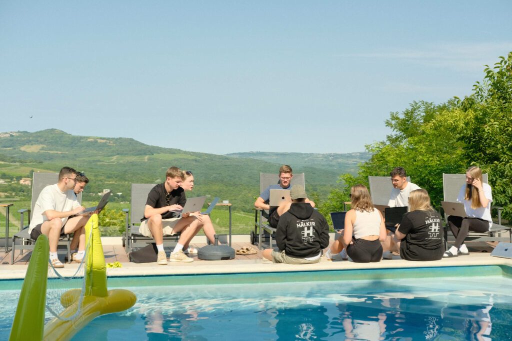 Junge Menschen arbeiten entspannt mit Laptops am Pool, umgeben von grüner Landschaft und sonnigem Himmel.