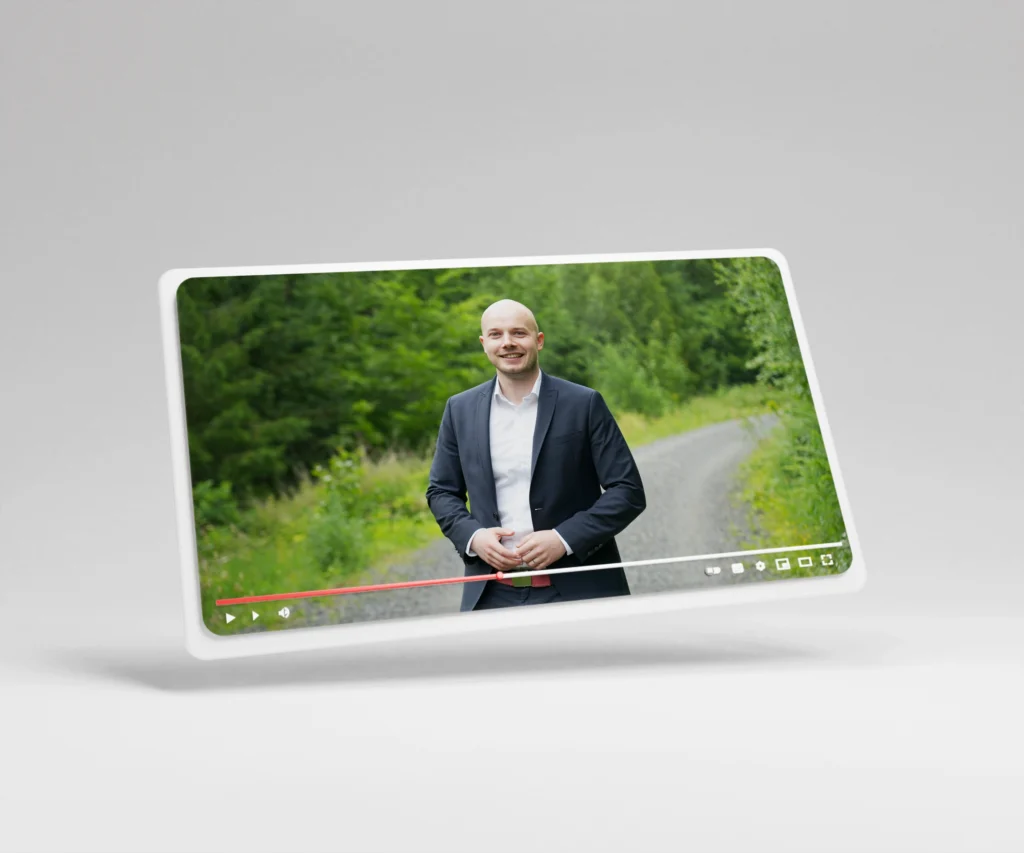 Ein Mann im Anzug steht lächelnd auf einem Schotterweg vor üppigem, grünem Laub und spricht.