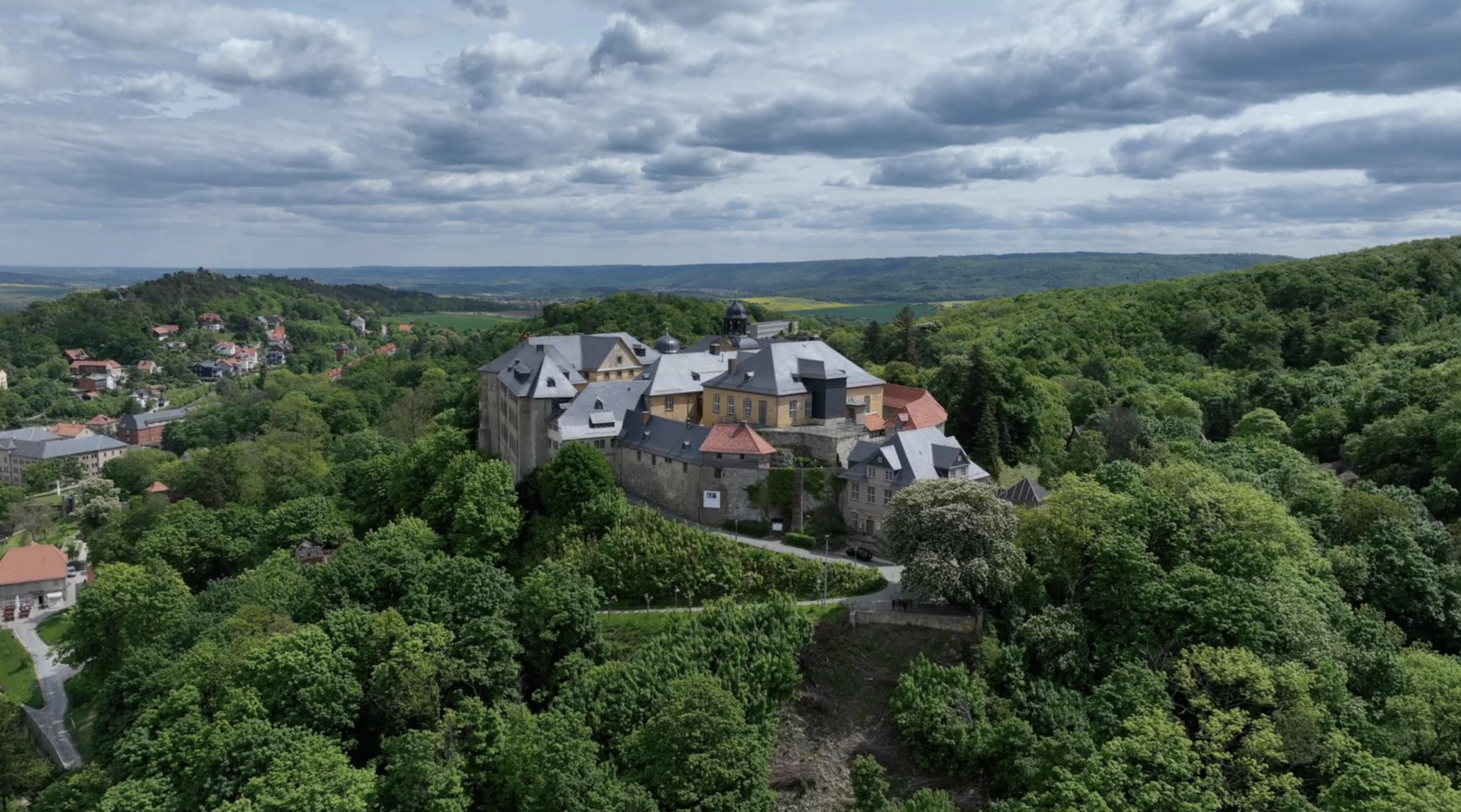Eine große Burg umgeben von dichtem Wald, unter einem wolkigen Himmel in einer idyllischen Landschaft.