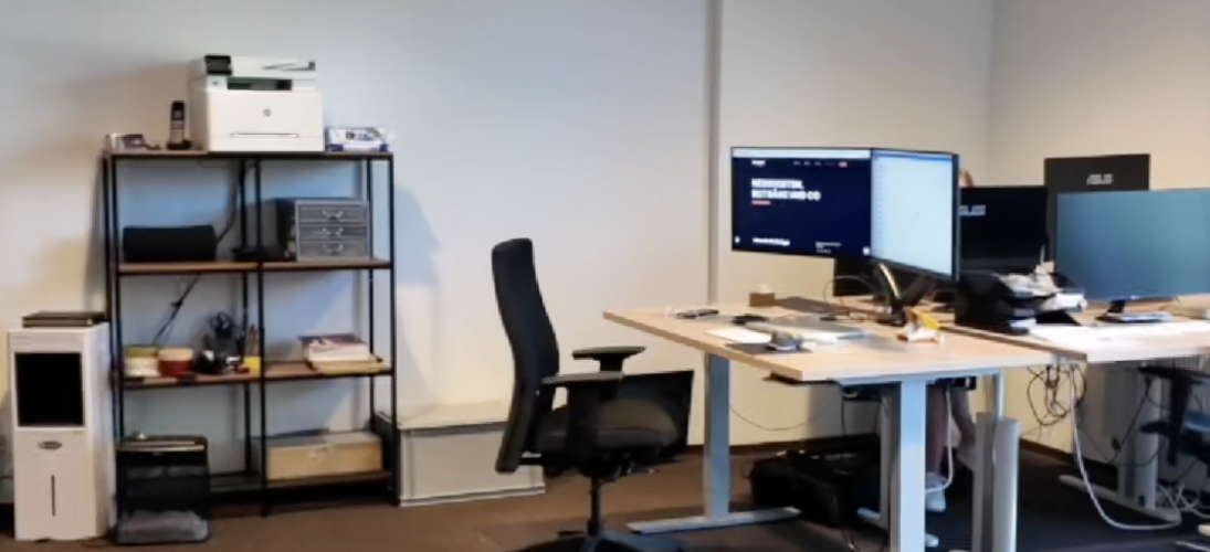 Modernes Büro mit Schreibtischen, Monitoren, ergonomischem Stuhl und Regal mit Büromaterial.