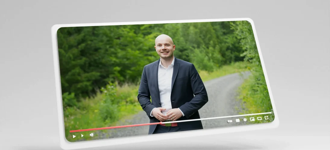 Ein Mann im Anzug steht lächelnd auf einem Schotterweg vor üppigem, grünem Laub und spricht.