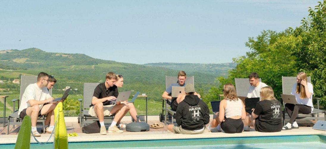 Junge Menschen arbeiten entspannt mit Laptops am Pool, umgeben von grüner Landschaft und sonnigem Himmel.