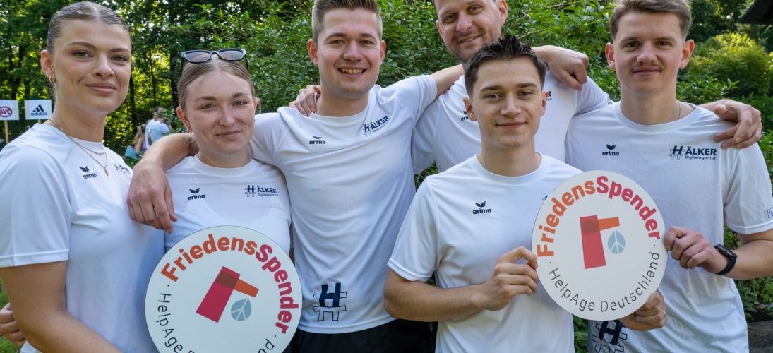 Sechs Personen in weißen Sportshirts halten Schilder von "FriedensSpender - HelpAge Deutschland" im Park.