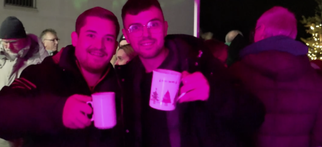 Zwei Personen stoßen unter einem roten Pavillon mit Tassen an, umgeben von geselligen Menschen.